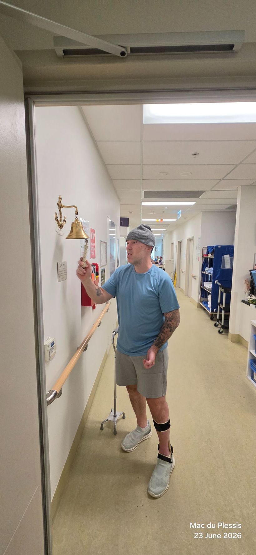 Man in a beanie ringing the end-of-treatment bell in a hospital corridor after finishing radiation therapy